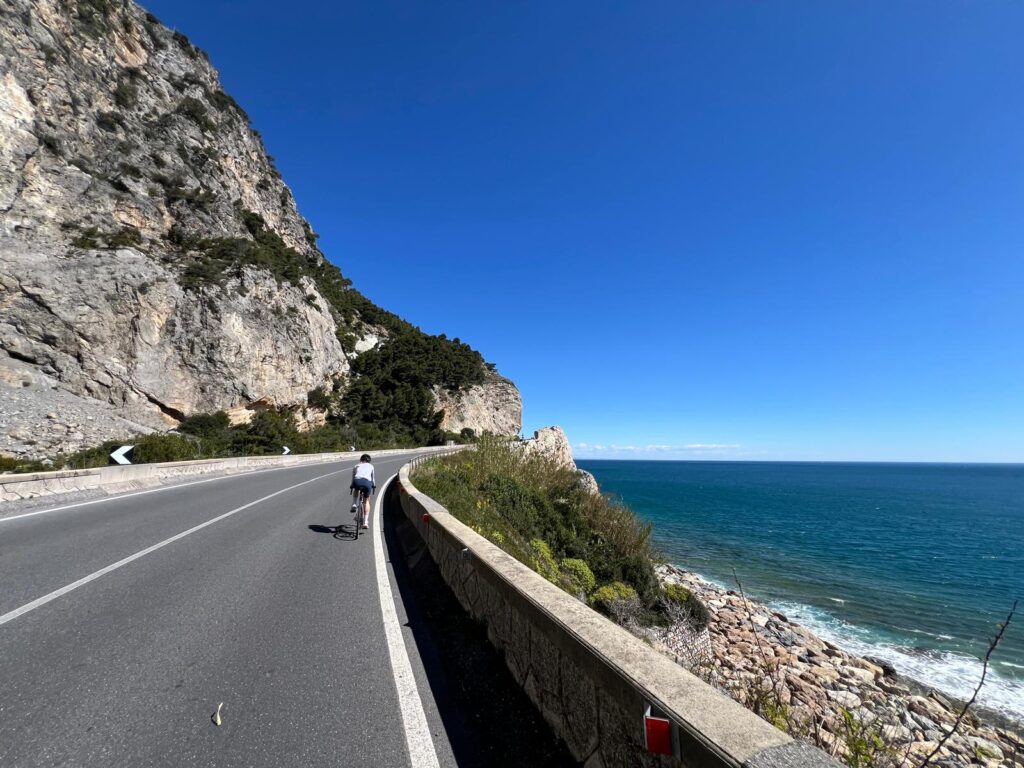 Küstenstraße in Ligurien direkt neben dem Meer. Auf der leeren Straße fahrt eine Radfahrerin mit einem Gravelbike.