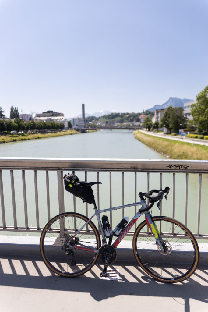 Start der Wossa Gravelbike-Tour in Salzburg. Foto mit Gravelbike auf einer Brücke und Salzburg im Hintergrund.