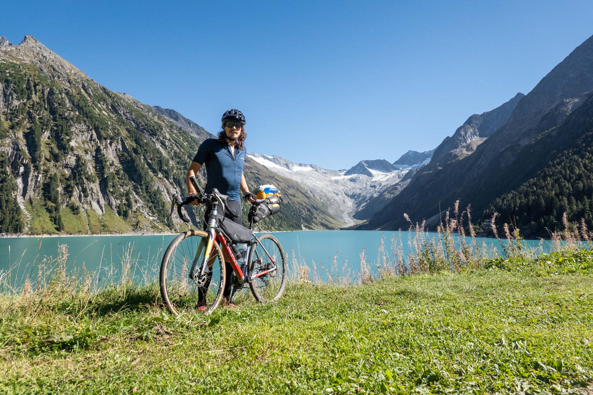 Ein Gravelbiker (Thomas Kargl) vor dem Schlegeisspeicher. Ein tiefblauer Speichersee im Zillertal mit hochalpiner Bergkette rundherum.